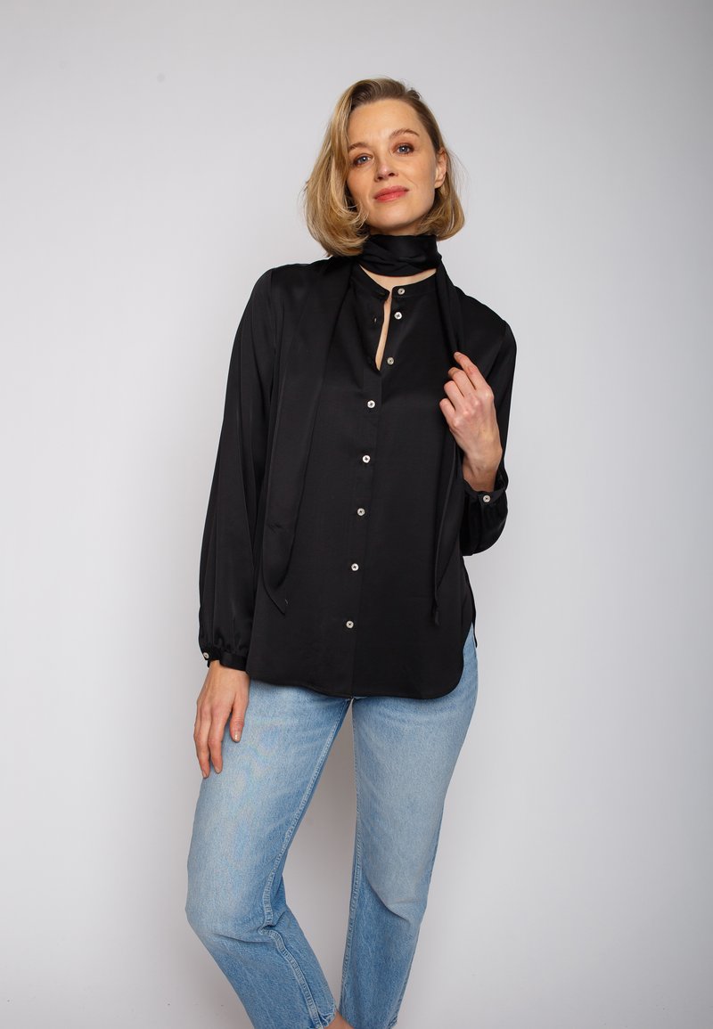 Czarna satynowa koszula na guziki z długimi rękawami, z detalem w postaci wiązania przy szyi i srebrnymi guzikami, zestawiona z jasnoniebieskimi dżinsami. Gładka faktura.