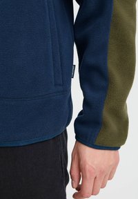 Veste en polaire avec un corps navy, des manches vert olive et un ourlet arrondi. Comprend une étiquette sur la couture latérale et une texture lisse.