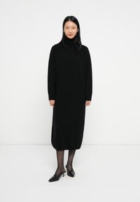 Moteris, apsirengusi juoda ilga rankovėmis turtleneck midi suknelę, permatomomis juodomis pėdkelnėmis ir juodais žemais kulniukais, stovi ant balto grindų.