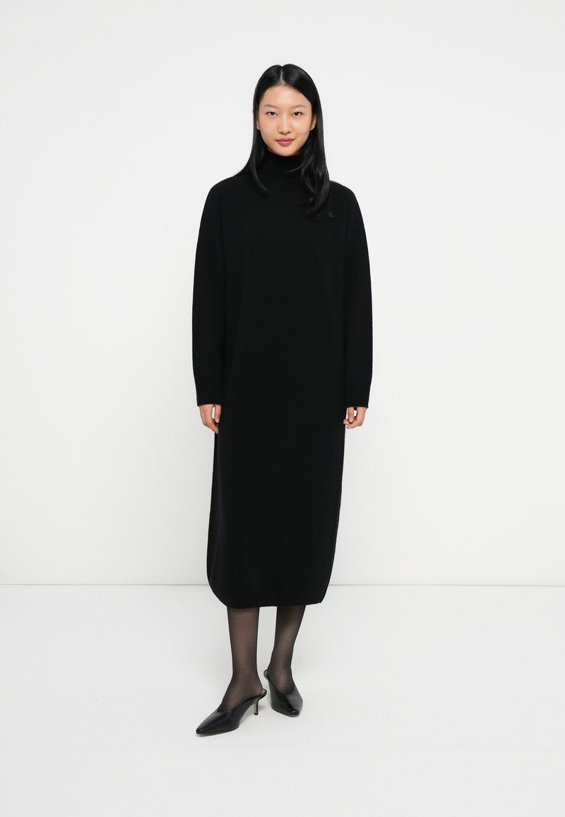 Moteris, apsirengusi juoda ilga rankovėmis turtleneck midi suknelę, permatomomis juodomis pėdkelnėmis ir juodais žemais kulniukais, stovi ant balto grindų.
