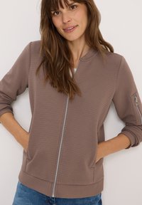 Getexturede bomberjack met rits in taupe, met ribbeldetails, zijzakken en een aansluitende tailleband. Voorzien van lange mouwen en een ronde halslijn.