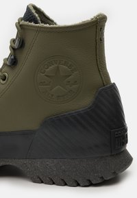 Khaki kožená teniska kotníkového střihu s texturovanou černou gumovou podrážkou, která má kulatou nášivku s logem a kontrastní švy podél límce.