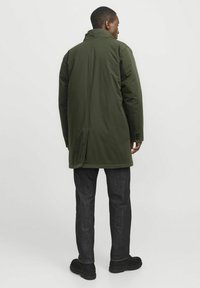 Cappotto verde imbottito con collo alto e spacco sul retro, caratterizzato da maniche lunghe e taglio dritto. Abbinato a jeans scuri slim-fit e calzature nere.