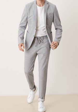 Uomo che indossa un blazer grigio chiaro e pantaloni abbinati con coulisse, sneakers bianche e una T-shirt bianca, che cammina in avanti su uno sfondo semplice.