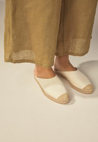 Vita slip-on skor med rund tå, juteaccent vid tån och sulan, uppvisade med beige, löst sittande linbyxor.