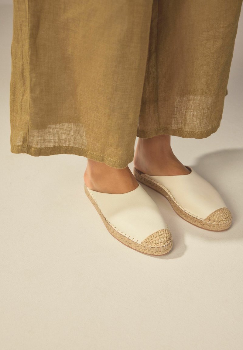 Vita slip-on skor med rund tå, juteaccent vid tån och sulan, uppvisade med beige, löst sittande linbyxor.
