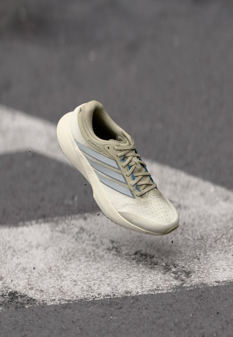 Lys beige løbesko med mesh-overdel, tre sølvstriber, blå accenter, tekstureret tåområde og polstret krave, vist mod en grå baggrund.