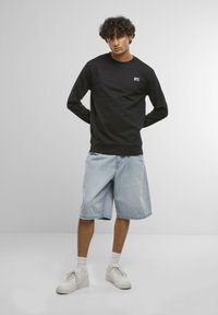 Felpa nera a maniche lunghe con logo "NYC", abbinata a pantaloni corti in denim chiaro e sneakers bianche. Lo sfondo è grigio neutro.