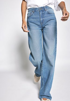 Personne portant un jean en denim bleu clair ample, un haut blanc et des chaussures beiges tissées à enfiler, marchant vers l'avant sur un fond uni.