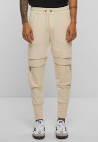 Beige joggebukser laget av myk stoff, med snor i midjen, store lommer og glidelåslukkinger ved lårene. Klassiske mansjetter ved anklene.