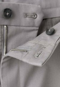 Primo piano di tessuto grigio con bottone, cerniera e dettagli di cucitura. Finitura strutturata e colore uniforme con accenti in metallo.