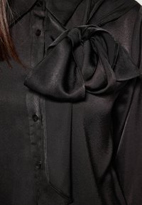 Blusa nera in satin con un grande fiocco drappeggiato sulla spalla sinistra. Presenta un collo a camicia e una chiusura con bottoni, texture liscia.