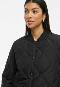 Νεαρή γυναίκα με ίσια μαύρα μαλλιά, φορώντας ένα μαύρο καπιτονέ μπουφάν με στρογγυλό γιακά και ασημένια σκουλαρίκια κρίκους, κοιτάζοντας προς τα πλάγια.