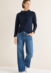 Kvinna iklädd en marinblå långärmad topp med dragsnören och vida blå jeans, stående med ena handen i fickan, tittande åt sidan.