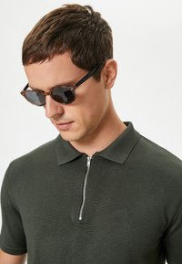Ciemnozielona koszulka polo z zamkiem błyskawicznym przy kołnierzu i teksturowanym materiałem. Model nosi brązowe okulary przeciwsłoneczne z czarnymi akcentami.