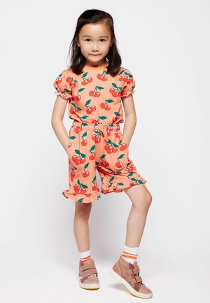 Mladé dievča stojí s rukami v nohaviciach, oblečené v broskyňovom outfit s červeným motívom čerešní, bielymi ponožkami a ružovými teniskami na suchý zips na bielom pozadí.