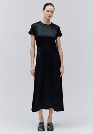 Žena stojaca v čiernych midi šatách s krátkym rukávom a čiernych slip-on topánkach s bielymi bodkami na jednoduchom svetlom pozadí.