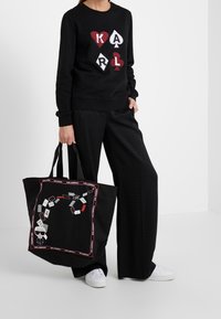 Svart sweatshirt med kortleksdesign och bokstäverna "KARL." Svarta mönstrade byxor. Stor svart väska med speltemattryck och accenter.