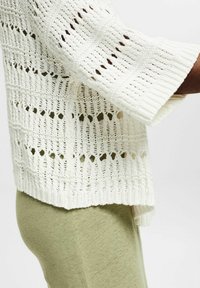 Suéter de punto en color crema con un patrón de calados y amplios puños de canalé. Combinado con pantalones ajustados de color verde claro, que muestran una tela texturizada.