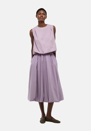 Femme debout en robe lilas sans manches avec taille froncée et jupe ample, portant des chaussures plates vert foncé, sur fond gris clair.