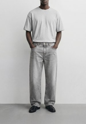 Άνδρας που φοράει ανοιχτό γκρι oversized μπλουζάκι, φαρδιά ξεβαμμένα γκρι τζιν, μαύρα μοκασίνια, στέκεται με τα χέρια στις τσέπες του μπροστά σε απλό λευκό φόντο.