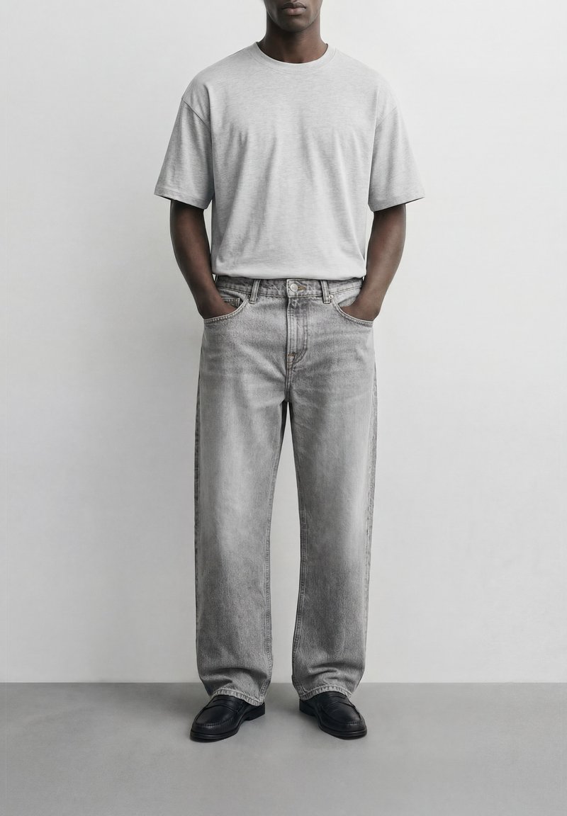 Άνδρας που φοράει ανοιχτό γκρι oversized μπλουζάκι, φαρδιά ξεβαμμένα γκρι τζιν, μαύρα μοκασίνια, στέκεται με τα χέρια στις τσέπες του μπροστά σε απλό λευκό φόντο.