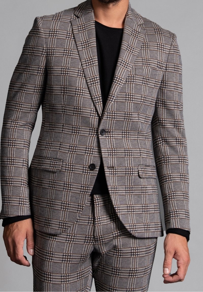 Homme portant un costume à carreaux marron et gris avec une chemise noire, debout devant un fond gris uni.