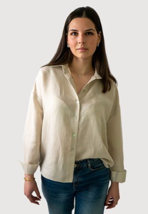 Femme aux longs cheveux foncés portant une chemise beige à boutons, un jean bleu, un bracelet en or et de petites boucles d'oreilles en anneau, debout devant un arrière-plan uni.