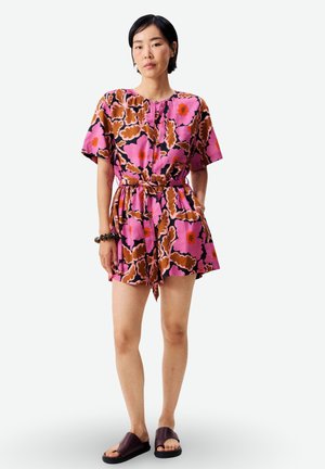 Femme portant une combinaison courte à manches courtes avec des motifs floraux roses, bruns et orange, taille nouée, et des sandales slides foncées, debout avec les mains dans les poches.