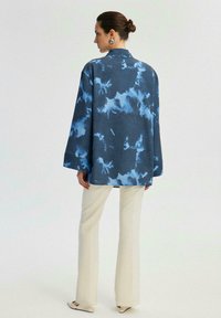 Camicia a maniche lunghe in tessuto blu scuro con un motivo astratto azzurro chiaro. Presenta un colletto e una vestibilità comoda, abbinata a pantaloni color crema.