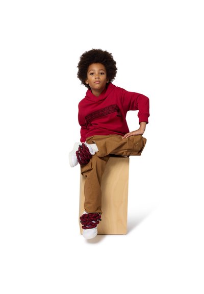 Czerwona bluza z kapturem o fakturowanym wzorze, zestawiona z beżowymi spodniami cargo. Sneakersy mają czerwone i granatowe akcenty. Siedzi na drewnianym blokowi.