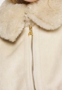 Giacca in shearling beige con colletto morbido, dotata di zip frontale e dettagli in metallo dorato; texture soffice e finitura liscia.