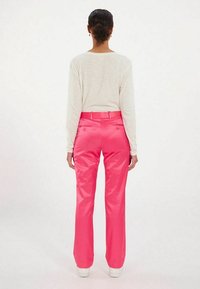 Pantalons satinés roses avec une coupe droite, présentant une texture lisse et des poches arrière, portés avec un haut léger à manches longues.