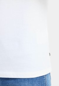 Hvid bomulds t-shirt med lige snit, korte ærmer og et lille mærkelabel syet på sidesømmen. Blå denim jeans synlige i bunden.