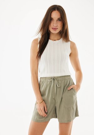 Młoda kobieta o długich brązowych włosach nosi bezrękawnik w białym kolorze i oliwkowe szorty z sznurkiem, pozując z jedną ręką w kieszeni.
