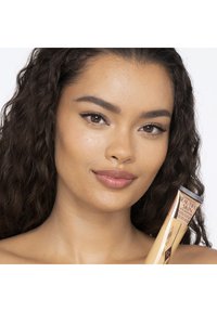 Woman with curly dark hair holds Charlotte's Beautiful Skin foundation tube, smiling with natural makeup and clear skin against a white background.