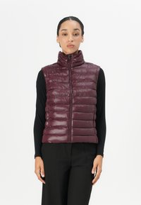 Bordeaux polstret vest med høy krage, med glidelås foran, quiltet design og et reflekterende logoaccent. Båret over en svart topp.