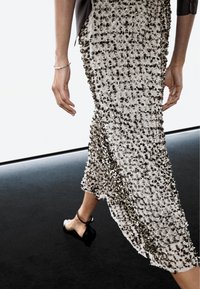 Persoon die loopt in een lange, getextureerde beige rok met zwarte versieringen, gezwarte enkellaarsjes met bandje en een zilveren armband om de linkerpols.