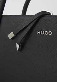 Melnas ādas rokassoma ar teksturētu virsmu, sudraba aparatūras akcentiem un zīmolu "HUGO" logo. Aprīkota ar regulējamām ādas siksnām.