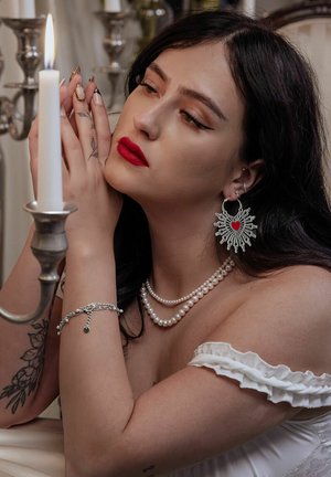 Perlenketten, Silberarmband und filigrane silberne Ohrringe mit rotem Herz. Das Model trägt ein schulterfreies, weißes Oberteil mit Rüschensäumen.