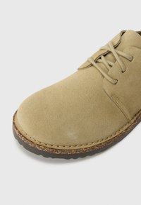 Scarpa in suede beige con punta arrotondata, lacci tradizionali e suola in gomma marrone texturizzata. Presenta cuciture visibili lungo il bordo.