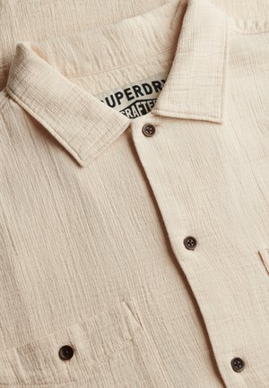 Camicia beige con trama, a bottoni, con colletto, tasca frontale visibile e bottoni scuri, che mostra parte dell'etichetta del marchio in bianco e nero all'interno.