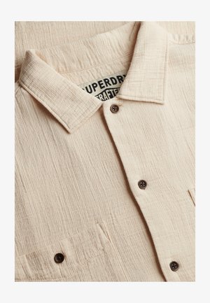Camicia beige con trama, a bottoni, con colletto, tasca frontale visibile e bottoni scuri, che mostra parte dell'etichetta del marchio in bianco e nero all'interno.