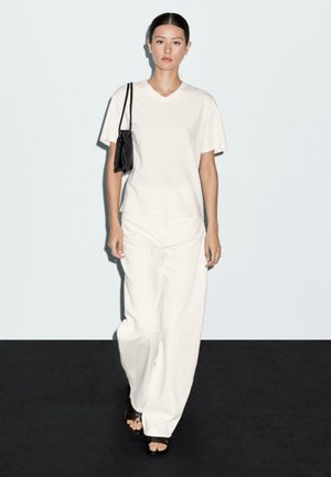 Junge Frau trägt weißes V-Ausschnitt-T-Shirt und weite weiße Hose, trägt schwarze Umhängetasche, geht auf einem schwarz-weißen Boden.