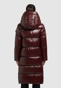 Hosszú, fényes bordó pufidzseki nagy kapucnival, vízszintes steppelt mintával és fekete logóval a háton, oldalán zsebfedő díszítéssel.