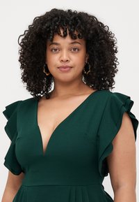 Robe vert foncé avec un décolleté en V profond, des manches volantées et des détails plissés, fabriquée à partir d'un tissu doux et lisse.
