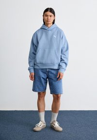Svijetloplava hoodie s kenguru džepom, bijelim logotipom, u kombinaciji s plavim traper šortsevima, sivim čarapama i bež tenisicama. Jednostavan dizajn s ležernim krojem.