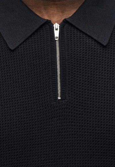 Κοντινό πλάνο ενός μαύρου πλεκτού πουκάμισου με ασημένιο ημισφαιρικό φερμουάρ και φαρδιά γιακά που φοράει ένα άτομο.