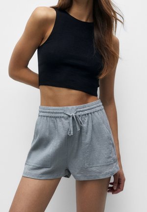 Femme portant un crop top noir sans manches et un short gris clair avec cordon de serrage sur un fond uni clair.