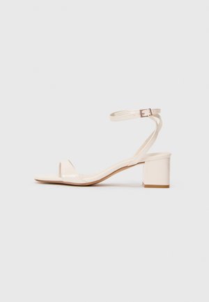 Beige patent leather sandal with a block heel, open toe, thin ankle strap, and minimalist design. Smooth texture with subtle shine.